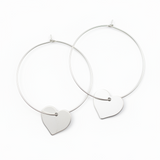 A pair of silver hoop earrings with heart charms attached, displayed against a dark background.