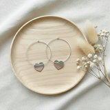 Silver hoop earrings with heart charms on a wooden plate with dried flowers.