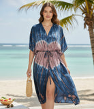 Woman in a tie-dye dress standing on a beach with palm trees and ocean in the background.
