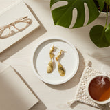 Gold earrings on a white plate with a cup of tea, glasses, and a plant in the background.