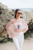 Model wearing a Suzie Blue Canada baby pink sheer mesh poncho over white pants on a beach with rocky cliffs