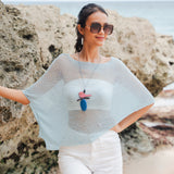 Model wearing a mint sheer mesh poncho by Suzie Blue Canada, styled with white pants and a bird pendant on a beach.