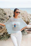 Model wearing a mint Suzie Blue Canada sheer mesh poncho with a bird pendant necklace on a beach by rocky cliffs.